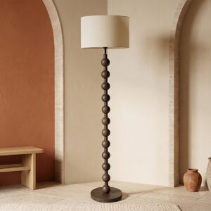 Un lampadaire vintage sculptural d’inspiration années 50 en bois foncé, avec une longue base à boules et un grand abat-jour clair, posé dans une pièce beige au style chaleureux.