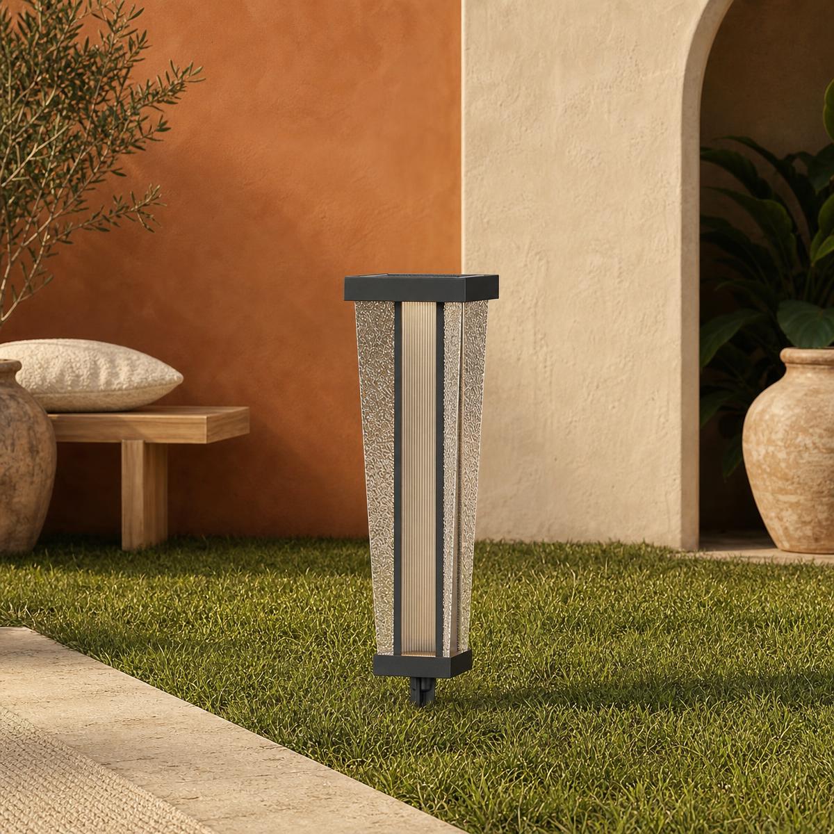 Un lampadaire extérieur LED design pour allée de jardin posé dans l’herbe, près d’une terrasse, avec une forme moderne et des côtés texturés.